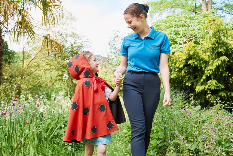 A Norland Nanny student holding hands with a girl wearing a cape in the Botanical Gardens, Bath